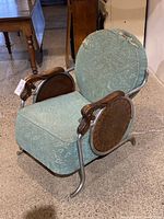 Side angle of art deco chair showing metal frame, wooden carved armrest, circular side panels, and worn teal upholstery.