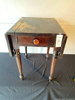 Front view of antique walnut drop leaf table with worn finish and visible patina, showing the single drawer with wooden knob and turned legs.
