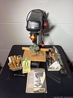 Front view of General International drill press mounted on a black stand with wooden platform and a plastic disc. Wooden accessories, Allen keys, metal hardware, and manual visible on the table.