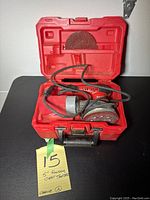 Red plastic carrying case with Milwaukee 5 inch random orbit sander inside, several sanding strips attached and loose.