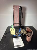 Front view of the vertical belt sander with sanding belt installed, extra sanding belt next to it, and instruction manual on the table.