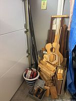 Overall view of scrap wood and metal pieces stacked against a wall, includes wooden boards, parts resembling guitar shapes, and some metal rods.