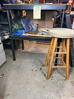 Black plastic two-level work shelf with peg board on side, assorted items on and under the shelf, wooden stool with fabric seat in front.