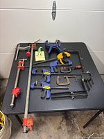 Overview of assorted clamps including metal pipe clamps, Irwin Quick-Grip clamps, DeWalt clamp, black metal clamps, and brackets on black table.