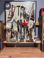 Wide view of pegboard with assorted hand tools hanging including saws, pliers, wrenches, scrapers, and measuring funnels.