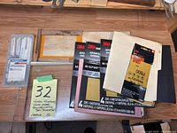 View of all lot items including reciprocating saw blades case, two wooden work surfaces, and multiple sheets of sandpaper from 3M and Washington brands.