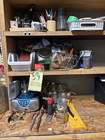 Photo of wooden shelves filled with various office and crafting supplies including pens, pencils, scissors, paintbrushes, lighters, jars with coins, small storage containers, and a Sylvania SRCD243 radio/CD player with broken antenna.