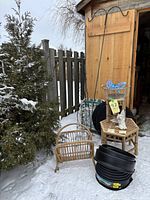 Image showing multiple garden items including rattan magazine rack, small lawn chair, metal trellis, shepherd's hooks, rattan tables, wire basket organizer, all sitting outdoors in snow next to wooden shed.