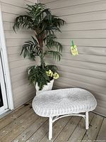 Artificial palm tree in white metal pot alongside white wicker table on a wooden porch floor.