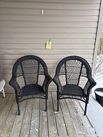 Two black wicker deck chairs placed side by side on a wooden deck with a light-colored wall behind them. A label with '46' is attached above the chairs.