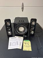 Front view of speaker system showing central subwoofer and two satellite speakers with instruction manuals laid out.