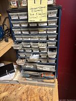 Blue metal storage organizer with labeled drawers full of assorted hardware, showing drawer labels like 'button', 'wing nut', 'key hook', 'magnet', 'springs', 'glue', 'metric' and more.