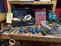 Front view of small plastic toolbox with various hand tools arranged in front, including screwdrivers with blue and red handles, pliers, adjustable wrench, a level, tape measures, sockets, wrench set, and a black pouch with RZ logo.