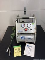 Front view of the California Air Tools air compressor showing handle, dual gauges (tank pressure and regulated pressure), red regulator knob, universal quick coupler, attached coiled hose, and owner's manual on a black surface.