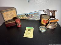 Wide view of antique items including the Tuckett's cigar box, Brill Brand baking powder tin, sharpener utensil, two glasses, an old iron, and the art saw with wooden handle.