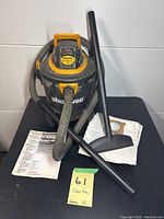 Front view of Shop-Vac vacuum with hose, wand, floor nozzle, instruction manual, and extra vacuum bags displayed on table.