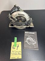 Full side view of Maximum 7 1/4 inch circular saw with black power cord wrapped around the saw. Includes handwritten note with lot number and item description and the instruction manual placed in front.