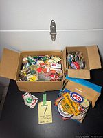 Photo of two open cardboard boxes containing various collectible match boxes, small pins, and patches, with some patches laid out beside the boxes