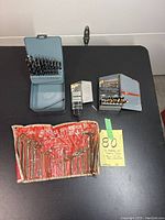 Overview of three metal cases with various sized drill bits and a red roll containing hex keys laid on black surface with a sticky note