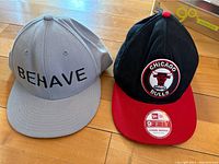 Two ball caps side by side. On the left, a grey 'Behave' cap. On the right, a black and red Chicago Bulls cap.
