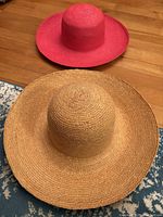Photo showing both sun hats: one natural straw colored floppy wide brim hat in front and one bright pink floppy wide brim hat behind on wood floor.