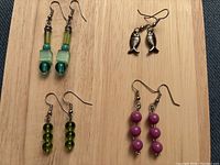 Four pairs of handmade earrings with green, purple beads and fish design shown on wooden background.