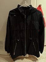 Front view of black Zara Basics women's jacket on hanger, showing full length, zipper closure, hood, and zippered pockets with cinching elastics at waist