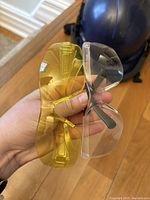 Hand holding two pairs of safety glasses, one clear lens and one yellow tinted lens, background shows a black hard hat on the floor.