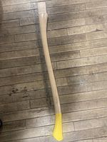 Full view of a wooden ax handle showing the natural wood finish and yellow-painted grip end on a wooden floor.