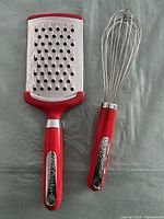 Photo of two Betty Crocker kitchen tools: a stainless steel box grater and a metal balloon whisk, both with red plastic handles featuring the Betty Crocker logo.