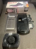 Black mandoline slicer with clear plastic slicing container and multiple blade attachments laid out alongside original box showing product branding and slices.