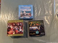 3 new puzzles with packaging issues, featuring various themes including a bird scene, Susan Brabeau indoor nostalgic scene, and a beach relaxation scene.