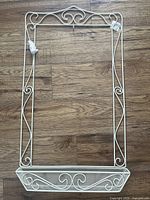 Front view of the new white wire calendar frame placed on wood flooring, showing its ornate scroll design and lower mesh basket.
