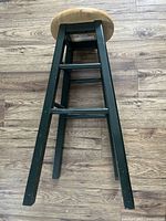 Side angle showing four dark painted legs, horizontal support bars, and round natural wood seat.