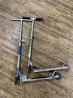 Photo shows two silver kick scooters with black handle grips placed side by side on a wooden floor. One scooter has a visible sticker 'Not So Amateur Amateurs.'