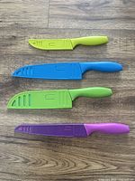 Top view of four knives with protective covers in light green, blue, green, and purple resting on wood floor.