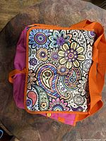 Front of tote bag showing colorful floral and paisley print panel with orange straps and pink body.