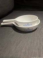 Side view of the two white ceramic Corning Ware pans stacked, showing the blue floral pattern on the sides and long handles.