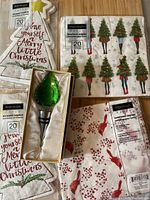 Overview of four packs of Harman Christmas napkins with varied festive designs and the boxed bottle stopper on wooden floor