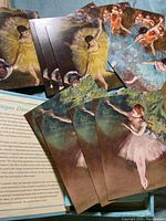 Multiple postcards with Edgar Degas ballerinas in yellow, blue and pink tutus spread on a surface with partial view of descriptive text.