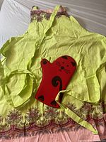 Photo showing entire lime green apron with pink floral and paisley trim along bottom and one strap visible with red cat-shaped hot pad placed on it.