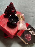 Small dark red Laughing Buddha statue on black holder with packet of incense sticks and booklet on box