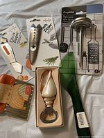 Kitchen tools arranged on white fabric including wavy knife, citrus zester, zero zipper herb strippeds, green spreader, garlic peeler/bottle opener, and Comphage Epicure meat thermometer set