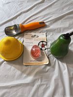 Lot items including orange handled metal scoop, yellow lemon-shaped juicer, heat sensitive egg timer in packaging, and green pear-shaped item on white background.