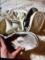 Top and sole view of silver snakeskin-textured ballet flats beside beige dust bag with Kruzers label.