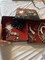 Photo of the opened jewelry box showing multiple pieces of jewelry inside, including necklaces, bracelets, a watch, a ring, and a black scrunchie, all inside two-tier lacquered box with floral design.