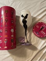 Wine glass with a black dress design on a white painted figure, standing on a purple base, alongside its original cylindrical box and lid showing various wine glass designs.