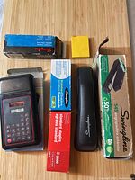 Photo showing all items laid out on a wooden surface, including stapler with box, three staple boxes, calculator, and a yellow sticky note pad