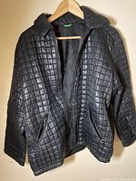 Full view of the black quilted jacket hanging on a wooden hanger showing the front with unzipped zipper and visible zipper pockets.