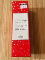 Front face of red rectangular box with small silver hearts and text stating 'Silicone Heart Tea Strainer' by Indigo with sale price sticker $8.00.